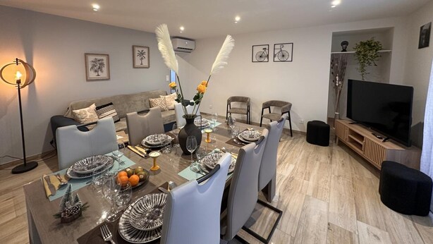 A modern living room with a dining area, featuring a sofa, chairs, a TV, and a dining table set with plates and glasses.