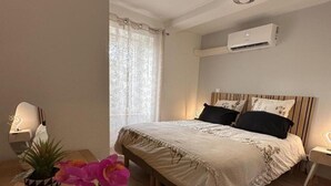 A bedroom with a bed, bedside tables, an air conditioning unit, a window with curtains, and a potted plant.