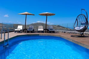 Ein Pool mit Liegestühlen und Sonnenschirmen, umgeben von einer Holzterrasse mit Blick auf eine gebirgige Landschaft.