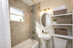 A bathroom with a bathtub, a sink, a mirror, and shelves with towels and plants. - Historic Uptown 2BD 1BA - 8 min to Downtown & Pier (St. Petersburg)