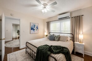 Une chambre à coucher comprenant un lit, des tables de chevet, un ventilateur de plafond, un appareil de climatisation, une fenêtre avec des rideaux et une porte donnant sur une autre pièce.