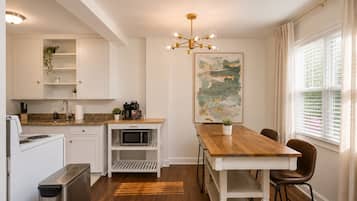Een moderne keuken met witte kasten, een houten eettafel en een groot raam met jaloezieën.