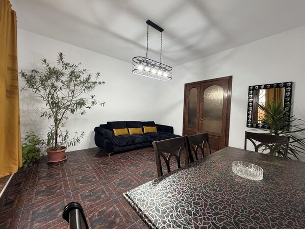 A dining area with a patterned table, a black sofa with gold cushions, a wooden door, and a potted plant.