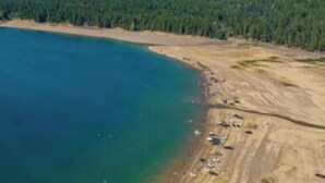Ett flygbildsbild av ett kustområde med ett klart, blått hav, en sandstrand och en skogbeklädd sluttning.