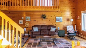 Interior acogedor de una cabaña con techo de madera, un sofá de cuero marrón, una mesa de centro de cristal y una escalera de madera.