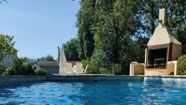 Une piscine aux eaux cristallines, bordée de carreaux, entourée de verdure et adossée à un bâtiment doté d’une cheminée.