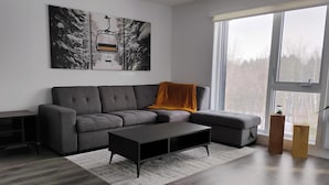 Ein modernes Wohnzimmer mit einem grauen Ecksofa, einem schwarzen Couchtisch und einem großen Fenster mit Blick auf Bäume.
