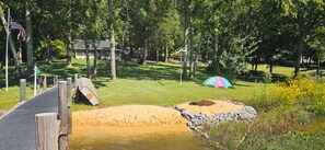 A park with a wooden bridge, a sandy area, a tent, and a house in the background.