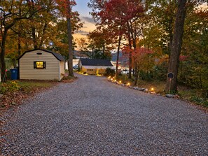 A gravel driveway leading to a small cabin with a lit window, surrounded by trees with autumn foliage.
