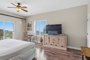 Une chambre à coucher avec un lit, un ventilateur de plafond, un téléviseur posé sur une commode en bois et un balcon avec vue.