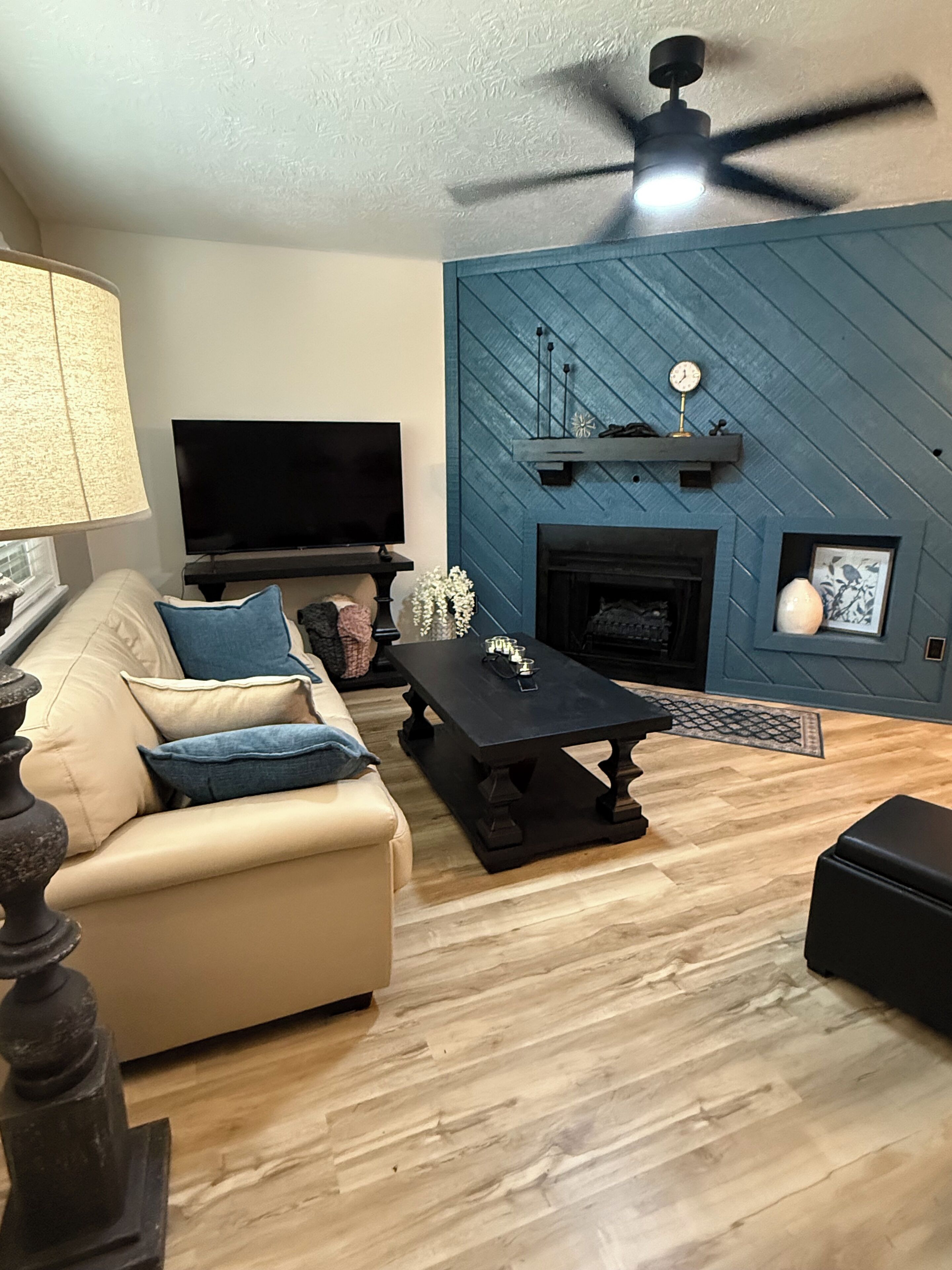 A living room with a beige sofa, a black coffee table, a flat-screen TV, a fireplace, and a ceiling fan.