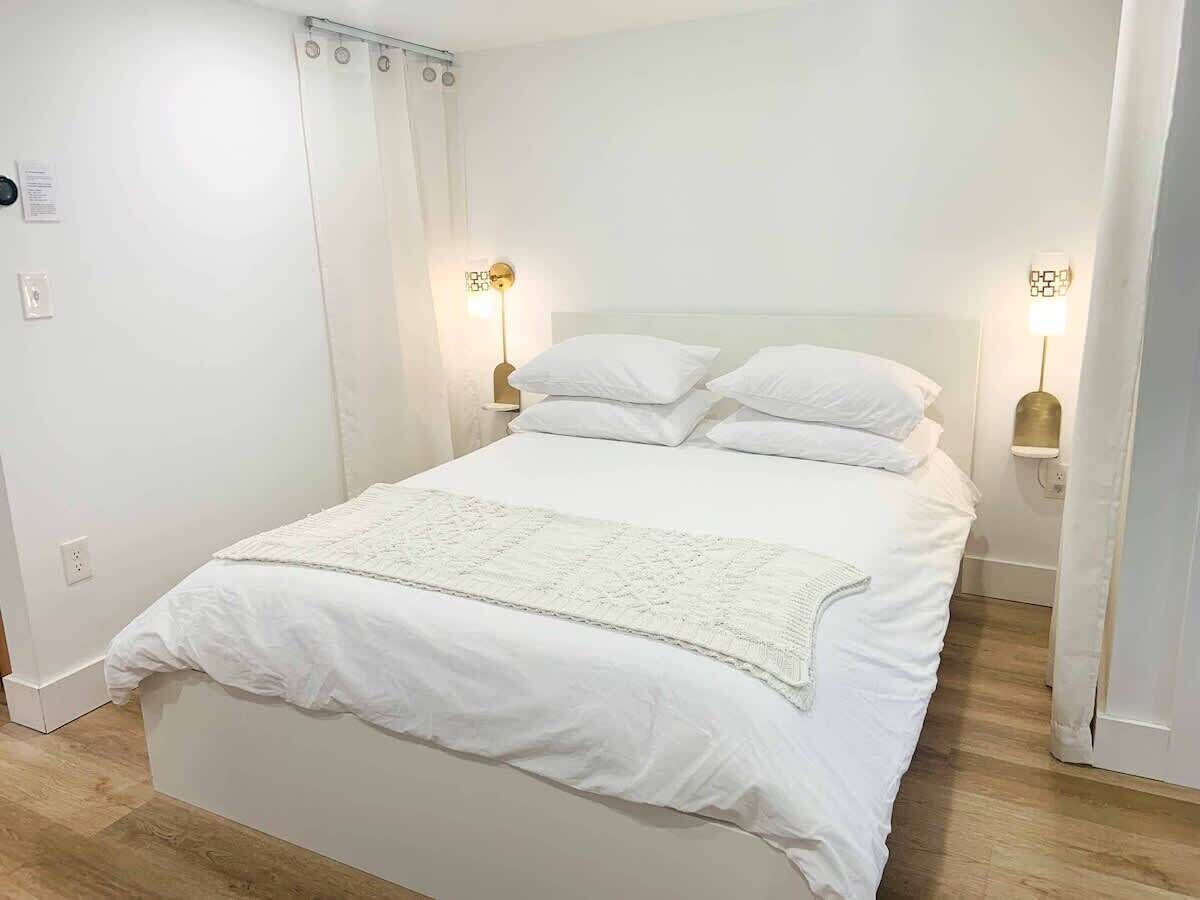 Una cama bien hecha con ropa de cama blanca y una manta de adorno blanca, dos lámparas de noche y un cabecero blanco.