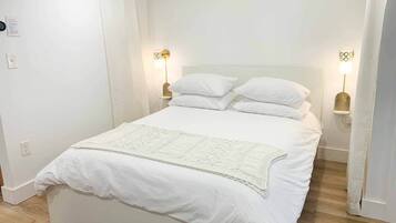 Una cama bien hecha con ropa de cama blanca y una manta de adorno blanca, dos lámparas de noche y un cabecero blanco.