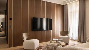 Una habitación de hotel moderna con un televisor de pantalla plana grande montado en la pared, una zona de estar confortable con dos sofás y una mesa de centro, y un ventanal con cortinas.