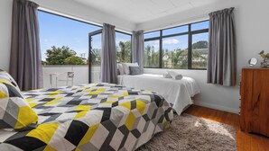 Ein modernes Schlafzimmer mit einem großen Bett, zwei Nachttischen, einer hölzernen Kommode und Blick auf Grün hinter großen Fenstern.