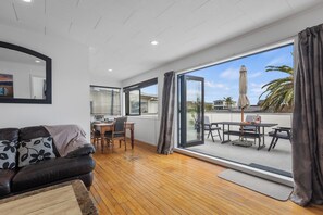 Una sala de estar moderna con piso de madera, un sofá de cuero, una zona de comedor con mesa y sillas, y un amplio ventanal corredizo que da a un patio exterior con mesa y sombrilla.