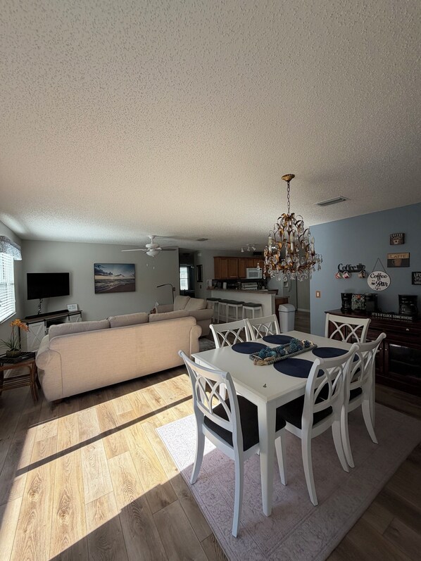 A spacious living room with a large sofa, dining table, and chairs, a kitchen area, and a ceiling fan. - Sumter landing Mallory Square (The Villages)