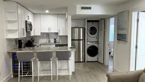 A modern kitchen with white cabinets, a stainless steel refrigerator, and a built-in washing machine. There are three bar stools and a beige sofa in the living area. - Naples Pier Condo 123 (Naples)