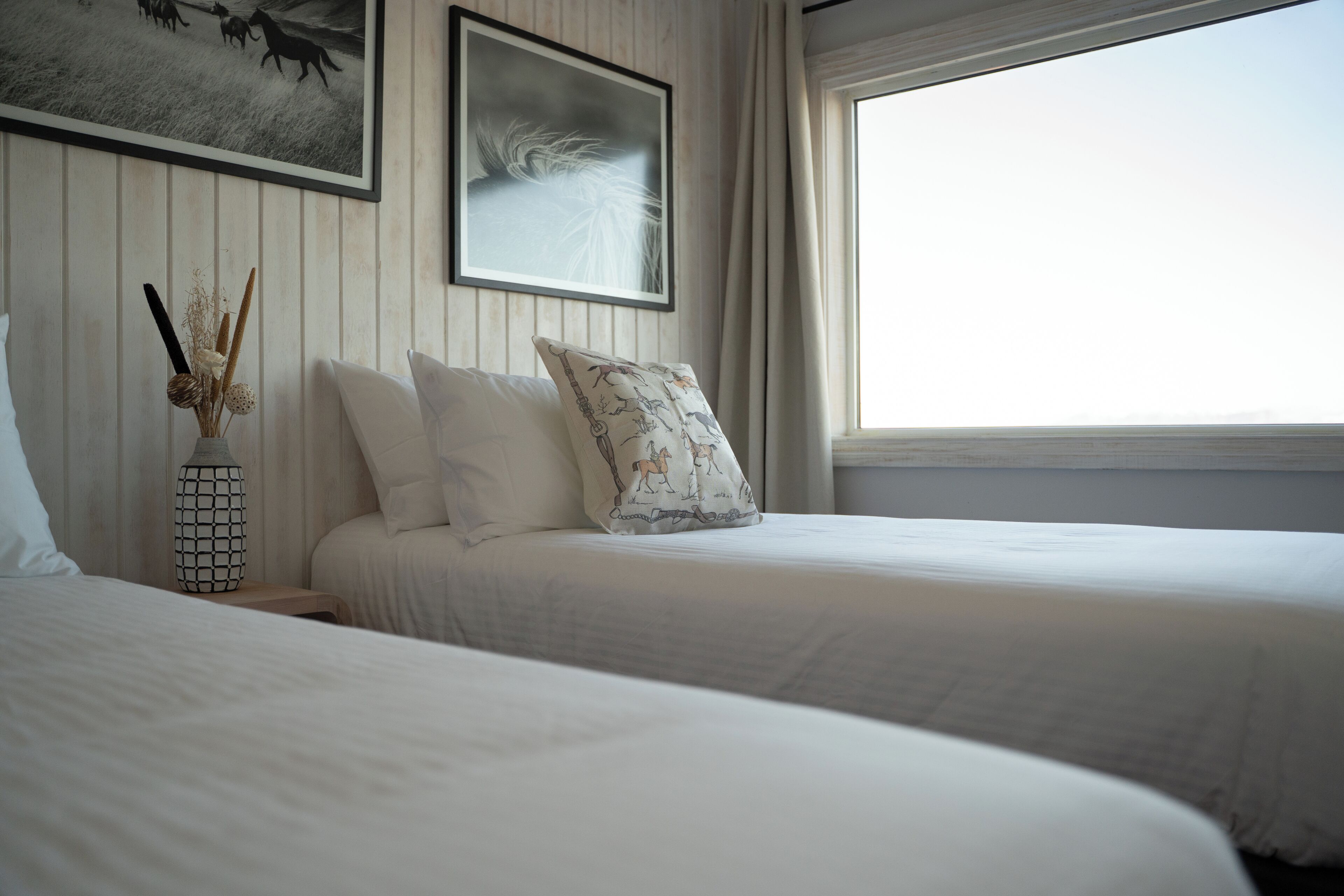 Een slaapkamer met twee bedden, een nachtkastje, een vaas met gedroogde planten en twee foto's in lijsten aan de muur.