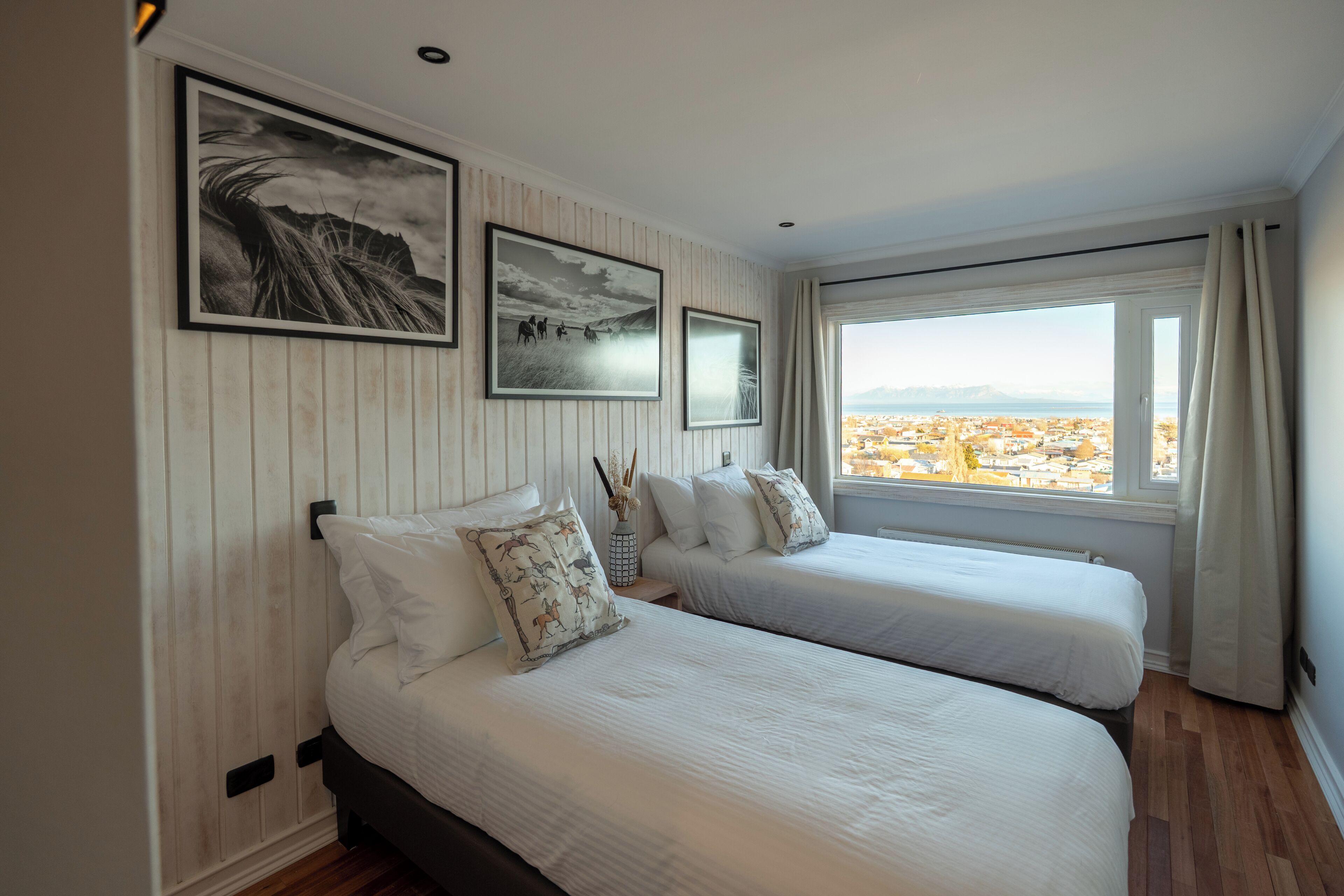 A bedroom with two beds, a large window with a view, and framed artwork on the wall.