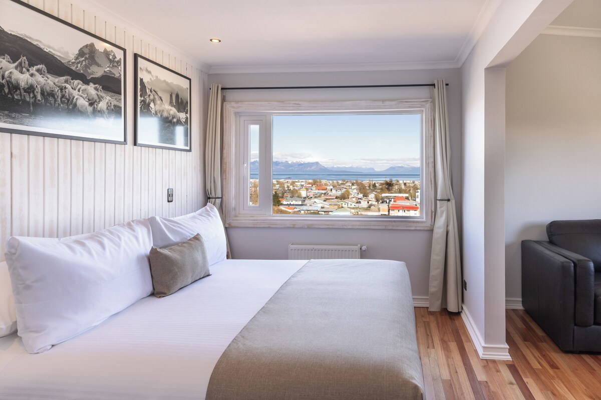 A hotel room with a bed, a view of a town, and a black leather sofa.