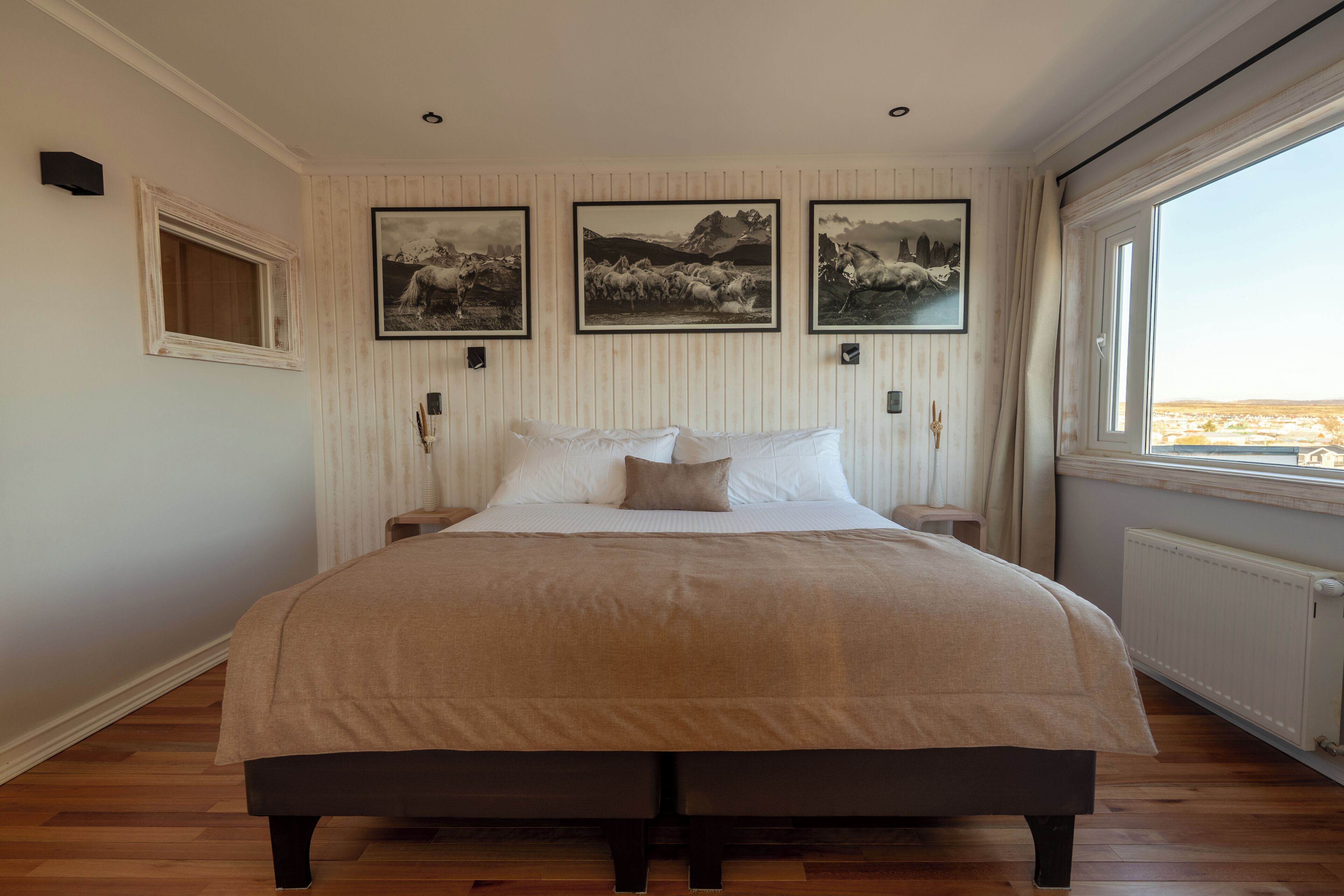 A bedroom with a large bed, three framed pictures on the wall, a window with curtains, and a radiator.