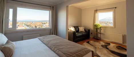 Un dormitorio con cama, un sofá de cuero negro, una planta en maceta y vistas a un paisaje urbano a través de grandes ventanales.