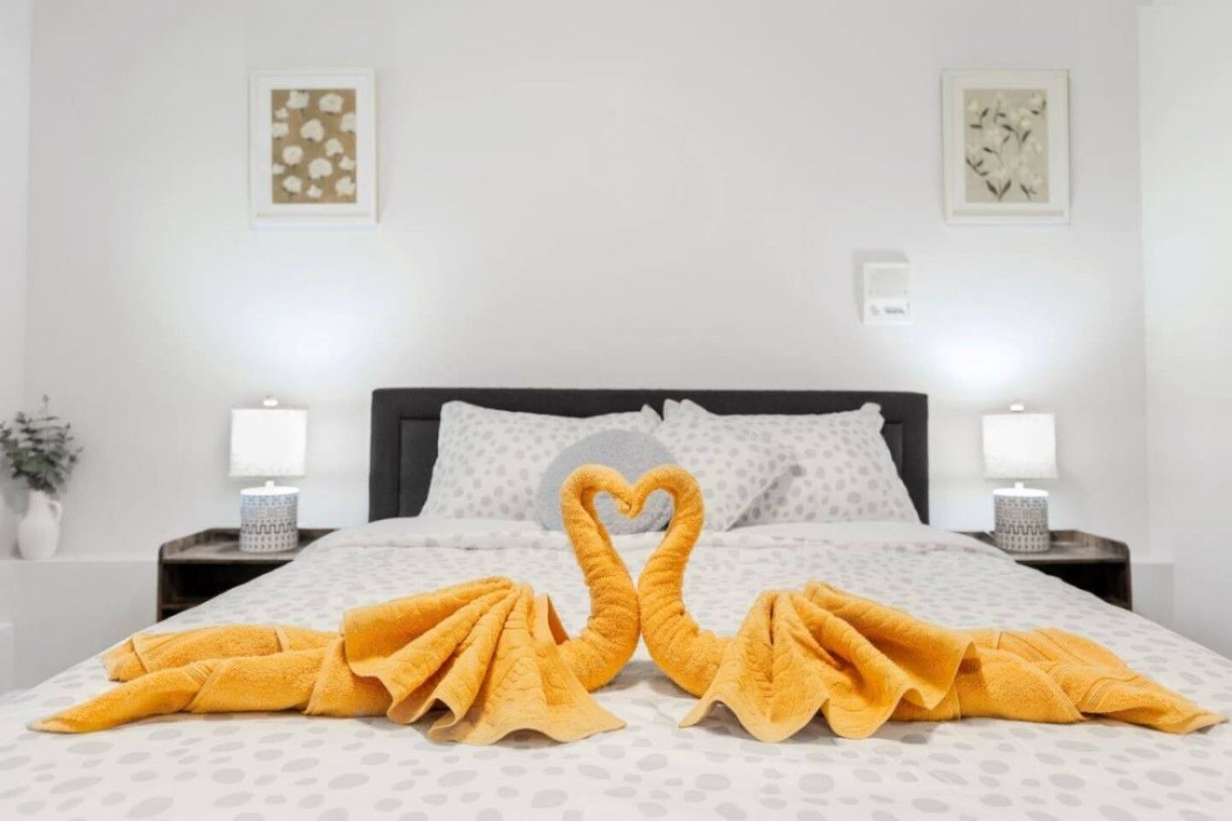 A hotel room with a bed, two nightstands, a lamp on each nightstand, and a heart-shaped towel arrangement on the bed.