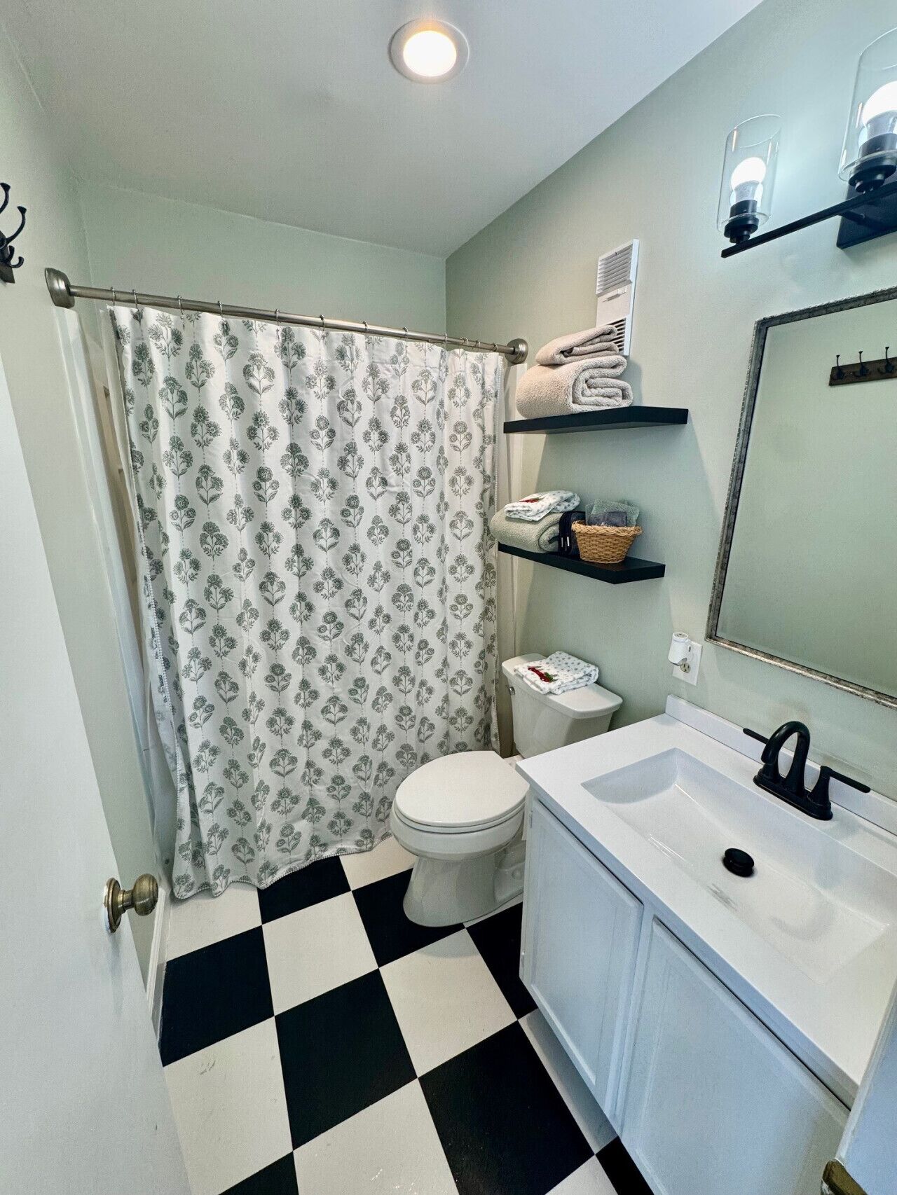 Ein Badezimmer mit Toilette und Waschbecken in Weiß, einem schwarz-weißen Schachfußboden, einem Duschvorhang mit Blumenmuster sowie Regalen mit Handtüchern und Körben.