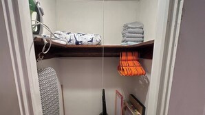 A small, narrow closet with a ceiling light, shelves for folded clothes, and a vacuum cleaner. - Simple Stay Steps from UF (Gainesville)