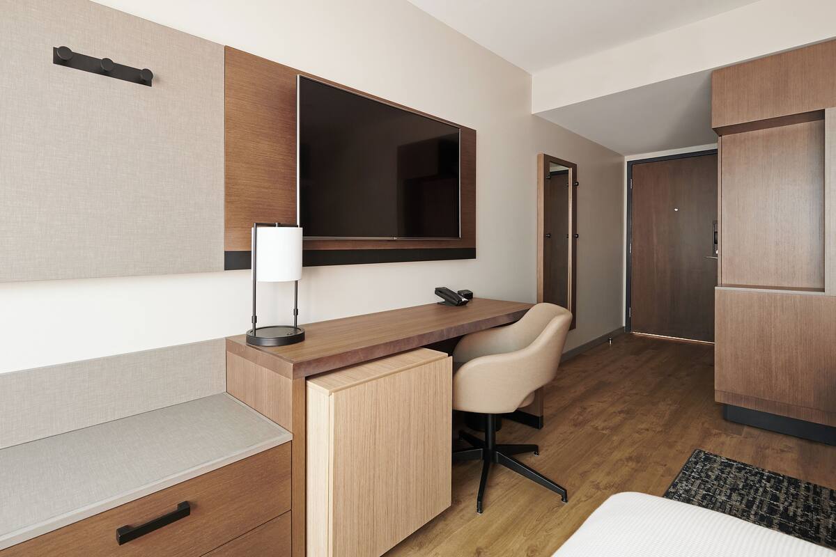 Een moderne hotelkamer met een houten bureau, een stoel, een nachtkastje, een lamp en een garderobe.