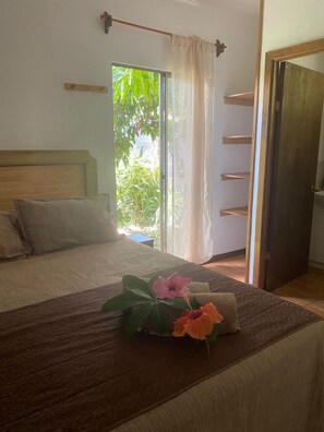 A bedroom with a bed, a wooden headboard, a window with curtains, and a view of greenery outside. - Pumakari Rapanui (Hanga Roa)