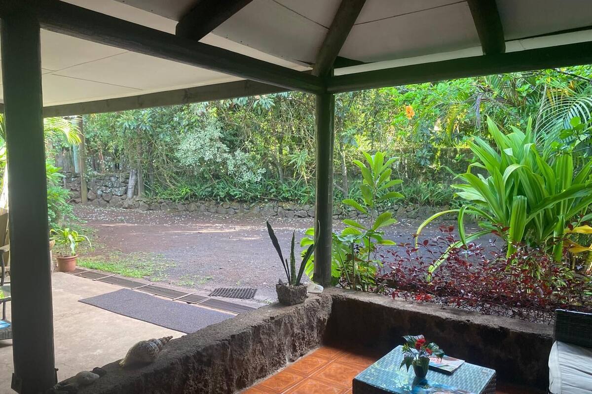 A covered outdoor area with a view of a garden, a paved path, and a seating area with a table and chairs.