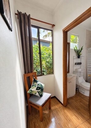 A room with a wooden chair, a window with curtains, a bathroom with a sink and toilet, and a view of greenery outside.
