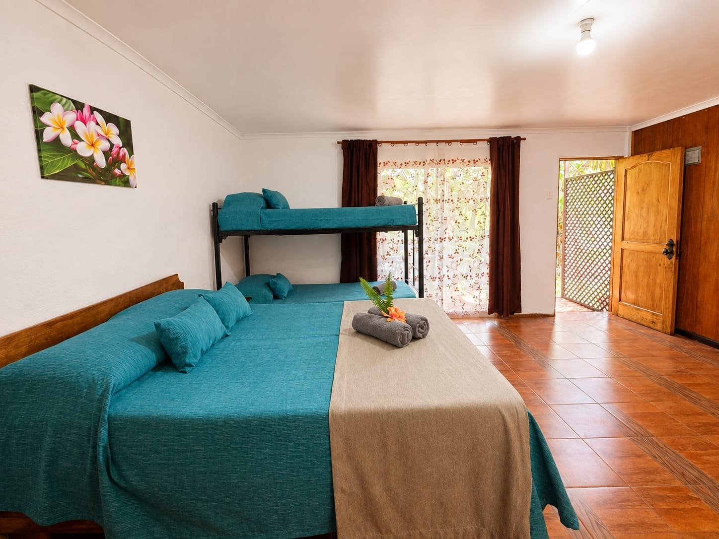A room with a bunk bed, a single bed, a table with a towel and a plant, and a wooden door.