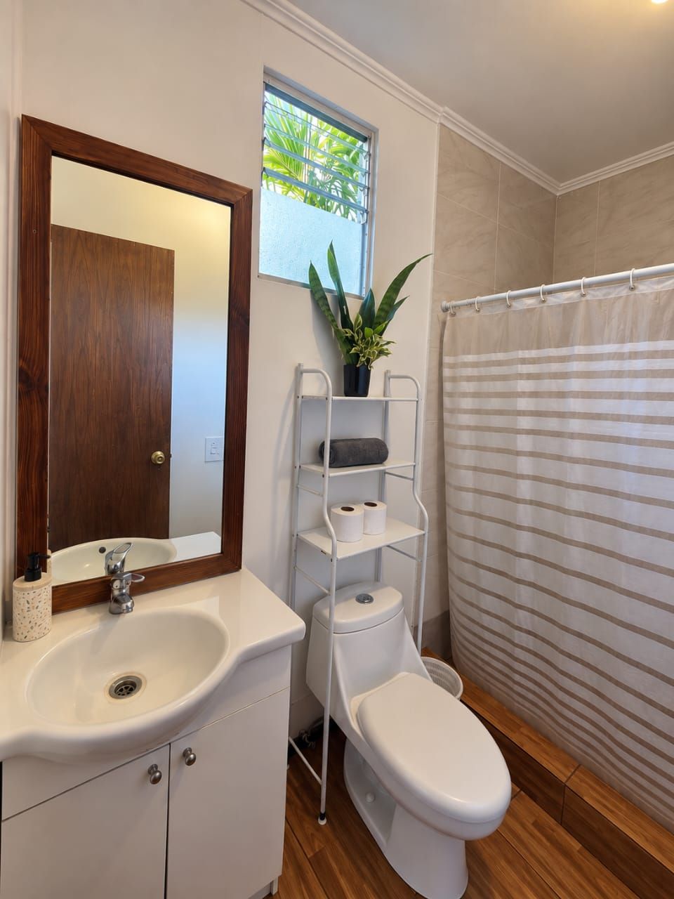 A bathroom with a white sink, a mirror with a wooden frame, a toilet, a shower curtain, and a potted plant.