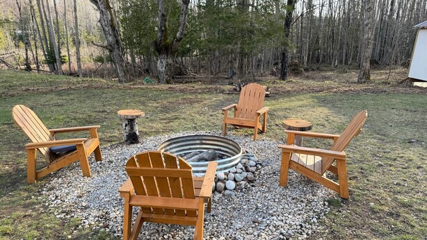 Een vuurkorf omringd door houten Adirondack-stoelen en een kleine tafel, gelegen in een grasrijke omgeving met bomen op de achtergrond.