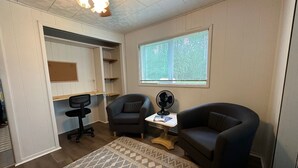 Een kamer met een bureau, twee fauteuils, een staande ventilator, een raam met jaloezieën en een plafondventilator.
