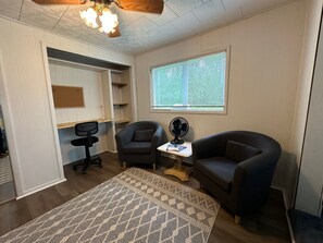 A room with a desk, two armchairs, a standing fan, a window with blinds, and a ceiling fan. - Charming 2-Bedroom Retreat in Curtis, Michigan (Germfask)