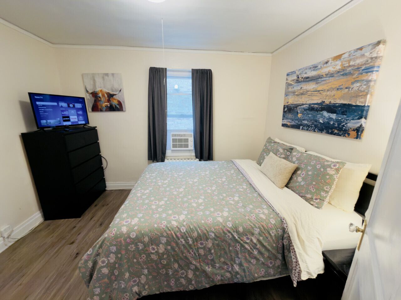 A bedroom with a bed, a dresser, a television, a window with curtains, and a painting on the wall.
