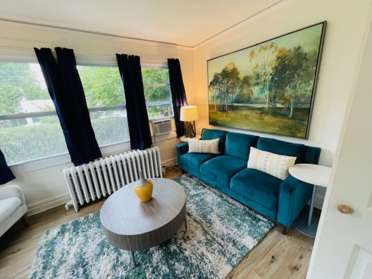 Un salon avec un canapé bleu, une table basse ronde, un tableau au mur et une fenêtre avec des rideaux.