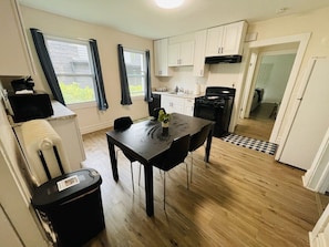 Cocina compacta con una mesa de comedor, un cubo de basura negro y una ventana con cortinas.