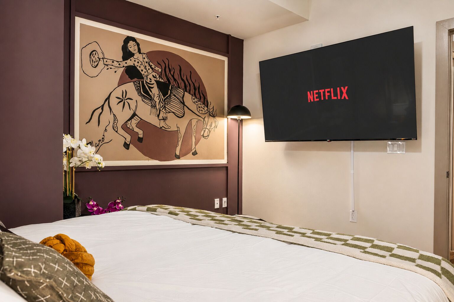 Een hotelkamer met een bed, een aan de muur bevestigde televisie waarop 'NETFLIX' wordt weergegeven, en een groot kunstwerk in een lijst boven het bed.