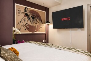 A hotel room with a bed, a wall-mounted TV displaying 'NETFLIX', and a large framed artwork above the bed.