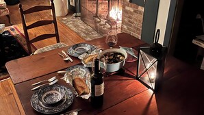 Una mesa de comedor preparada para dos, con platos, cubiertos, una botella de vino y un tazón de sopa. Se observa una chimenea con un marco de ladrillo y una repisa encima.