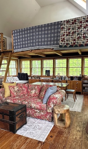 A cozy room with a loft bed, a floral sofa, a wooden chest, and a desk with books and a chalkboard.