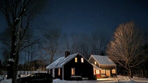 Una casa de dos pisos con tejado nevado y una chimenea, rodeada de árboles desnudos y un paisaje invernal.