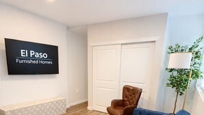 A furnished living room with a blue sofa, a wooden coffee table, a brown leather chair, and a television displaying "El Paso Furnished Homes". - Cozy Remodel 2 Story w/ King Bed / W/D / BBQ Patio (El Paso)