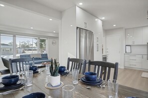 A modern dining area with a wooden table set for four, surrounded by chairs. A large window provides a view of the outdoors. - New 3BR Home - Minutes to the Falls! (Niagara Falls)