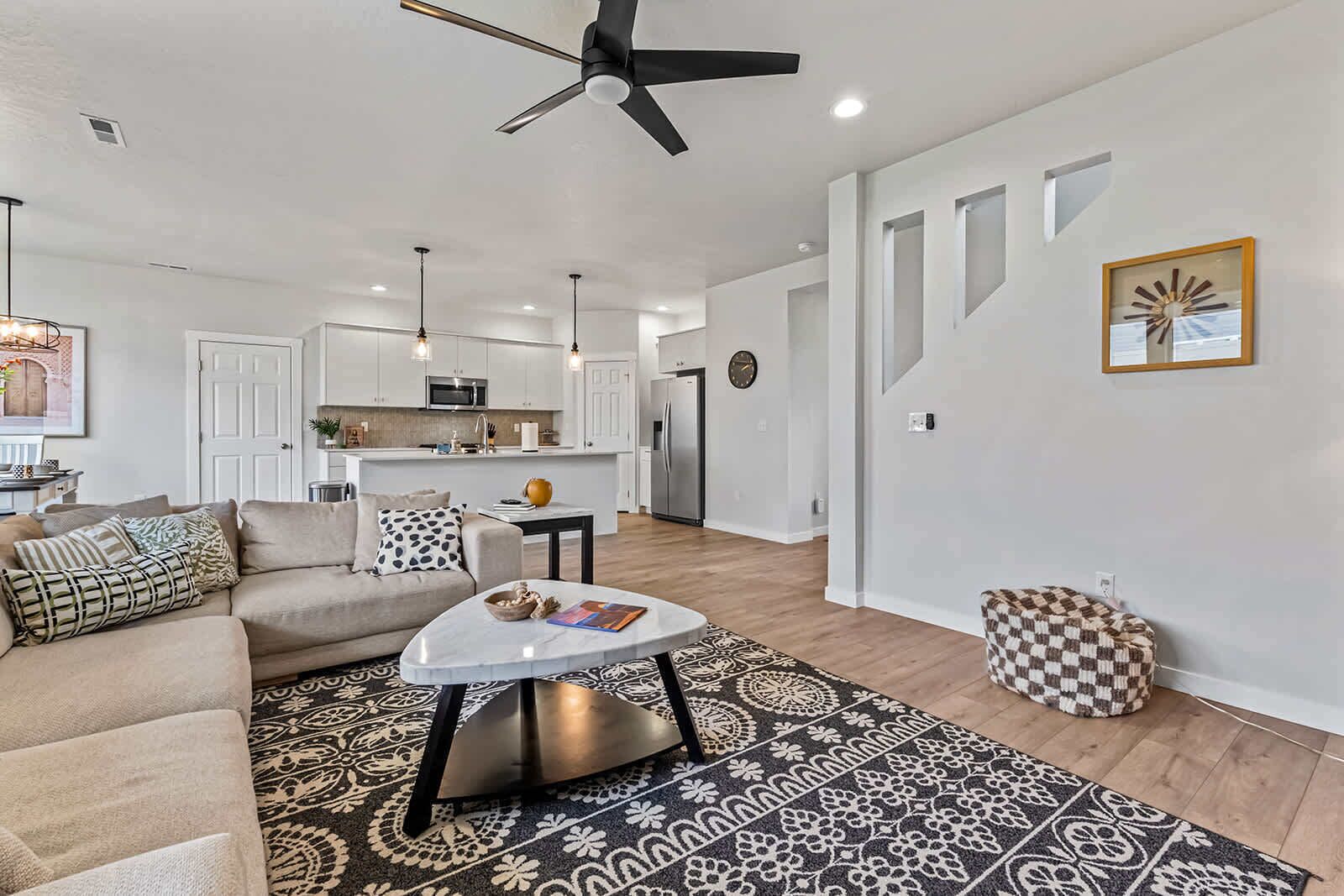 A modern living room with a sofa, coffee table, and patterned rug. A kitchen with white cabinets and a dining area is visible in the background.